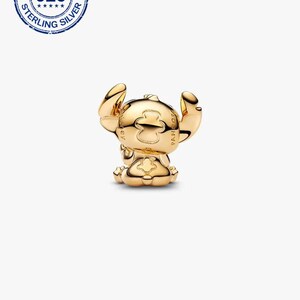 May include: A gold-coloured charm depicting a stylized figure with large ears in a seated position. The charm has a smooth, polished surface, likely made of metal. The charm is a collectible item.