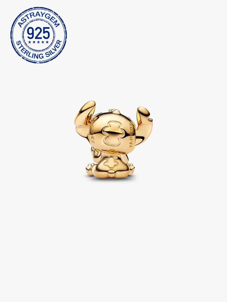 May include: A gold-coloured charm depicting a stylized figure with large ears in a seated position. The charm has a smooth, polished surface, likely made of metal. The charm is a collectible item.