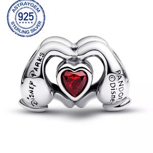 May include: A sterling silver charm with a heart-shaped red gemstone at the center. The charm features a design of two hands forming a heart shape. The words "Disney Parks" and "Pandora Disney" are inscribed on the charm.