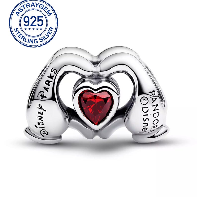May include: A sterling silver charm with a heart-shaped red gemstone at the center. The charm features a design of two hands forming a heart shape. The words "Disney Parks" and "Pandora Disney" are inscribed on the charm.