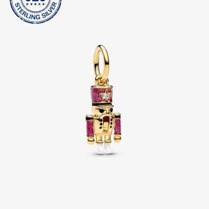 May include: A gold-colored charm in the shape of a toy soldier nutcracker. The charm has a red and glittery hat and arm cuffs, with a star-shaped gem on the hat. The charm has a gold loop at the top for attaching to a bracelet or necklace.