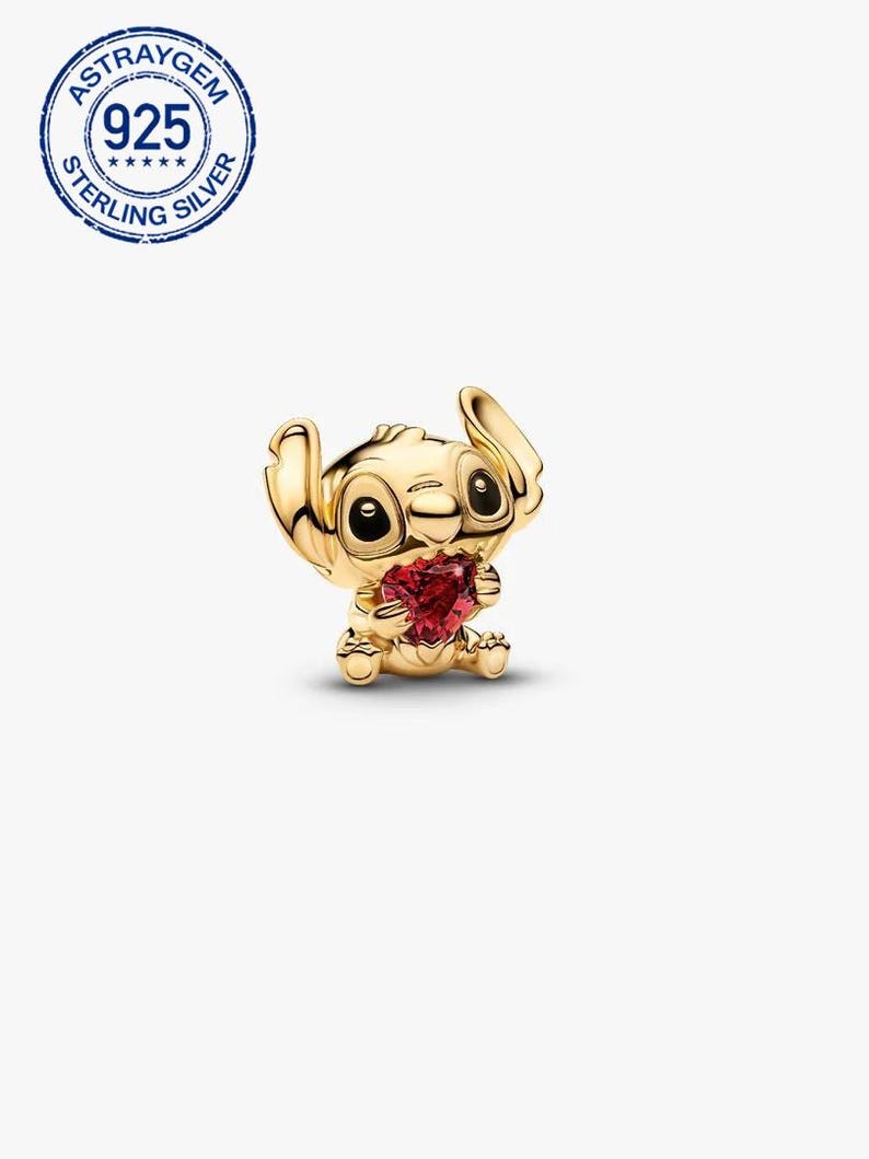 May include: A gold-coloured charm depicting a cartoon character holding a red heart-shaped gem. The charm has black eyes and a textured surface. The charm is stamped with "925 STERLING SILVER".