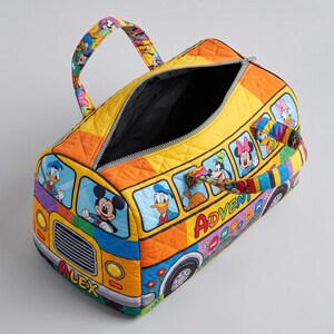Puede incluir: Una colorida bolsa de lona acolchada con un dise&ntilde;o de autob&uacute;s y personajes de Disney. La bolsa tiene un esquema de color amarillo, naranja y azul, con cierre de cremallera y asas. La palabra "ADVENTURE" es visible en el lateral.