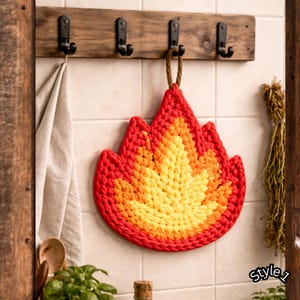 May include: A handmade, crochet trivet in the shape of a flame, featuring a gradient of red, orange, and yellow. The trivet is hanging on a wooden rack with black hooks. A beige towel hangs to the left.