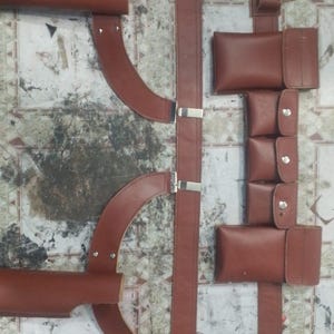 May include: Brown leather harness with multiple straps, pouches, and metal hardware. The harness features curved shoulder pieces and adjustable straps, suitable for various applications. The leather has a rich, reddish-brown hue.