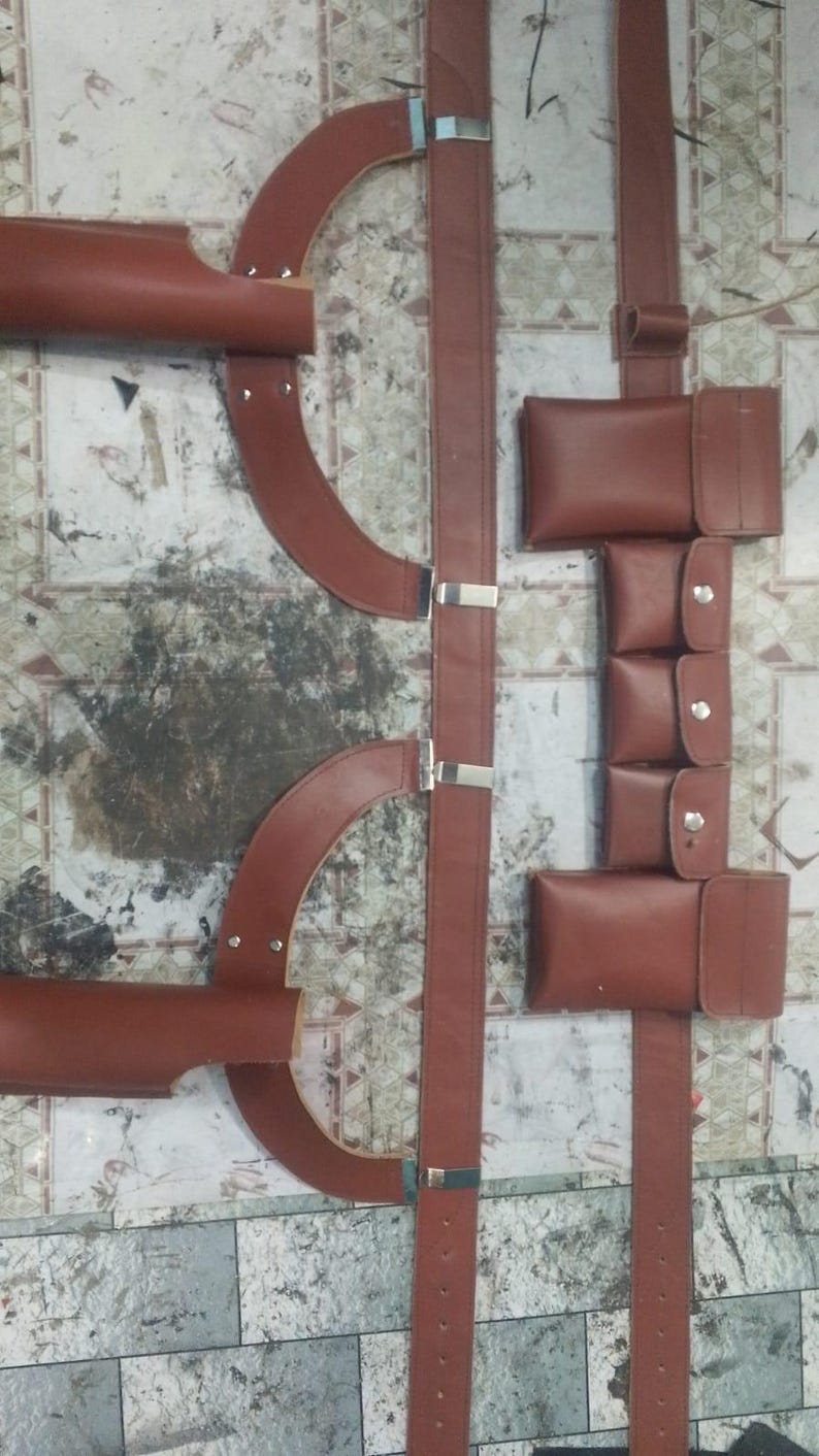 May include: Brown leather harness with multiple straps, pouches, and metal hardware. The harness features curved shoulder pieces and adjustable straps, suitable for various applications. The leather has a rich, reddish-brown hue.