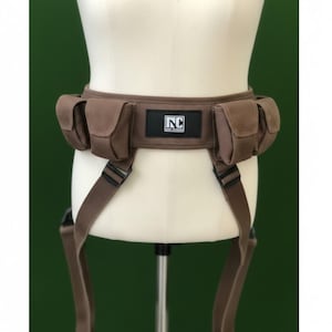 Könnte beinhalten: Ein brauner Utility-Gürtel mit vier aufgesetzten Taschen und verstellbaren Riemen mit schwarzen Kunststoffschnallen. Ein rechteckiger schwarzer Aufnäher ist vorne mittig angebracht, mit dem weißen Text NC und kleinerem Text darunter, auf einer Puppe.