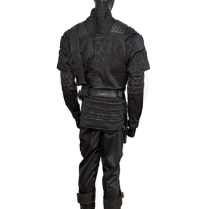 Pu&ograve; includere: Costume tattico nero con collo alto, spallacci e tasche multiple. L'outfit include giacca, pantaloni e cintura, il tutto in una finitura scura e opaca. Le maniche presentano un motivo geometrico.