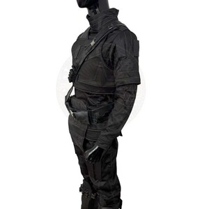 Pu&ograve; includere: Tuta tattica nera con collo alto, spalline e tasche multiple. L'outfit include un gilet, una cintura e guanti. Le maniche hanno cuciture geometriche. Il manichino &egrave; su sfondo bianco.