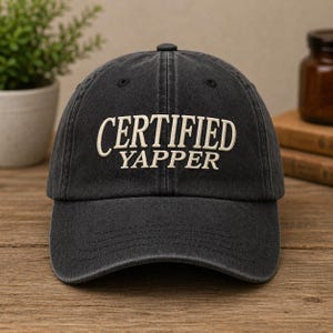 May include: A dark gray baseball cap with the embroidered text "CERTIFIED YAPPER" in white. The cap has a curved brim and a small button on top. The hat appears to be made of a soft, worn material.