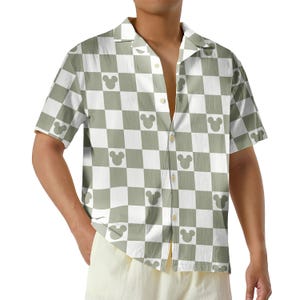May include: A short-sleeved button-down shirt featuring a checkerboard pattern in white and olive green. The design includes Mickey Mouse silhouettes within the squares. The shirt has a classic collar and is made of a lightweight fabric.