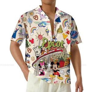 May include: Cream-colored button-down shirt with a colorful all-over print featuring Disney characters and landmarks. The shirt has short sleeves and a relaxed fit. The word "Epcot" is prominently displayed in green script.