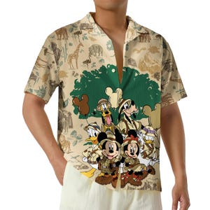 May include: A short-sleeved button-down shirt in a light tan color featuring a safari-themed print. The design includes illustrations of Mickey Mouse, Minnie Mouse, Donald Duck, Goofy, and Pluto in explorer gear, set against a backdrop of trees and animals.