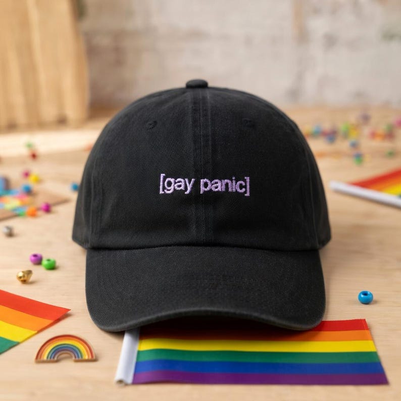 May include: Black baseball cap with the text "[gay panic]" embroidered in purple. The cap is displayed with a rainbow flag and other colorful accessories, suggesting an LGBTQ+ theme.