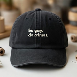 May include: Black baseball cap with the embroidered text "be gay, do crimes." The cap has a curved brim and a fabric strap closure. The hat is made of a soft, durable material.