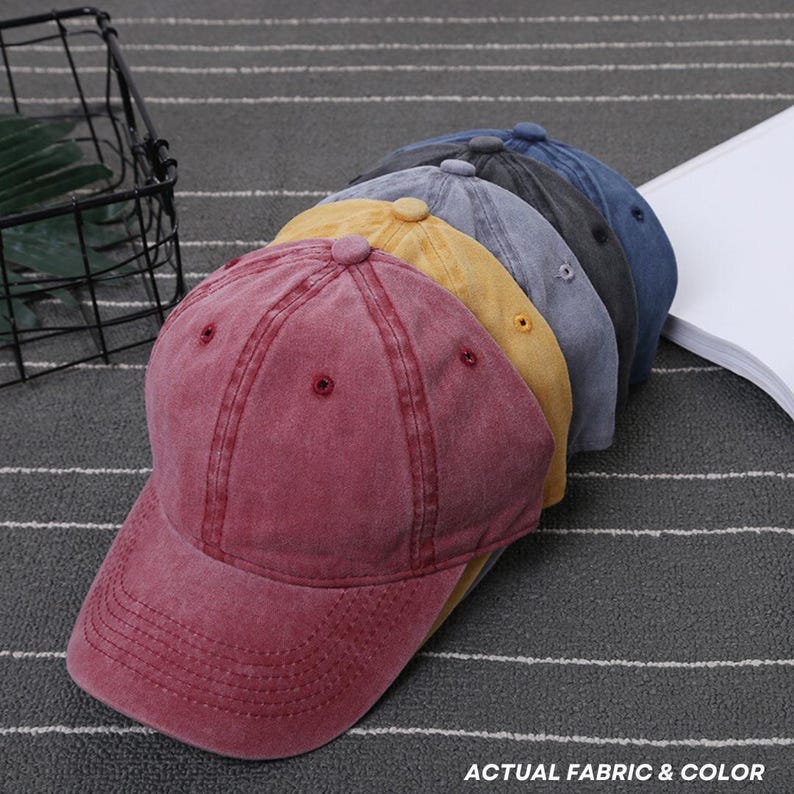 May include: A row of baseball caps in various colors: burgundy, yellow, gray, dark gray, and blue. The caps have a classic design with a curved brim and adjustable strap. The text "ACTUAL FABRIC & COLOR" is visible.