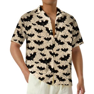 May include: A short-sleeved button-down shirt featuring a repeating pattern of black bats and thin grey spiderwebs against a light beige background. The shirt has a classic collar, a front button placket, and a relaxed fit with a straight hemline.