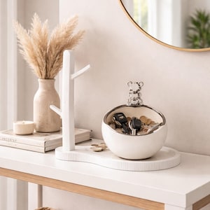 Puede incluir: Un llavero de cerámica blanca con borde plateado, que contiene llaves y monedas. Un colgador de llaves decorativo blanco en forma de árbol está al lado. Sobre una mesa blanca, un jarrón contiene flores secas.