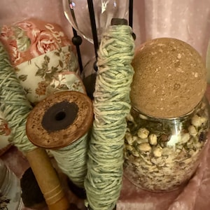May include: A collection of craft supplies, including spools of green yarn, a vintage wooden spool, and a glass jar filled with dried botanicals. A decorative hourglass and floral fabric add to the vintage aesthetic.