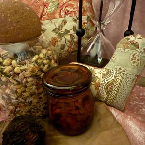 Puede incluir: Una variedad de objetos decorativos, incluyendo un tarro de cristal con tapa marrón, un reloj de arena transparente y un tarro de cristal lleno de semillas. El fondo presenta cojines con estampado floral y tela rosa.