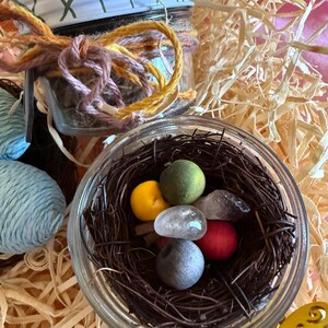 Puede incluir: Un tarro de cristal con tapa metálica, atado con hilo naranja, morado y amarillo. En el interior, un pequeño nido de pájaro hecho de ramitas contiene piedras lisas y coloridas. El tarro está rodeado de material de embalaje en forma de paja.