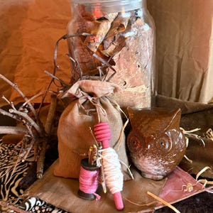 Op de afbeelding: Een glazen pot met een deksel gevuld met gedroogde bladeren, een kleine jute zak, een roze en witte spoelpop en een bruin uilbeeldje. De items zijn gerangschikt op een bruin oppervlak met zebraprint stof.