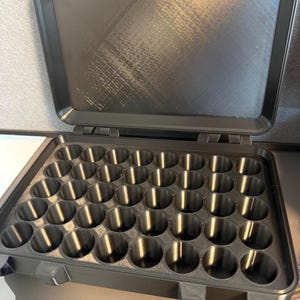 May include: A black plastic case with an open lid. The interior of the case has 35 cylindrical compartments. The case is designed to hold small items. The case is made of a durable plastic material.