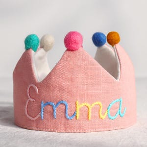 May include: A pink fabric crown with the name "Emma" embroidered in blue, yellow, and green thread. The crown is topped with colorful pom-poms in teal, white, pink, blue, and orange. A festive accessory.