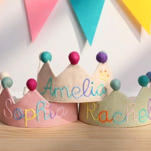 May include: Four fabric crowns with colorful pom-poms and embroidered names: Sophia, Amelia, and Rachel. The crowns are in shades of pink, beige, and green. Colorful triangle banners are in the background.
