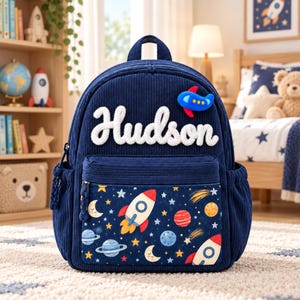 May include: A navy blue corduroy backpack featuring the name Hudson in white textured lettering. The front pocket displays a colourful space-themed pattern with rockets, planets, stars, and moons. A small blue aeroplane patch is attached above.