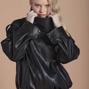 Puede incluir: Chaqueta de cuero negra con cuello alto abotonado y mangas largas. La chaqueta tiene un aspecto texturizado y una cintura entallada. La modelo lleva la chaqueta y tiene el pelo rubio.