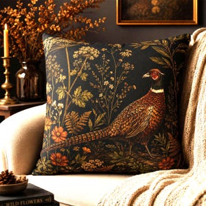 Op de afbeelding: Een vierkant sierkussen met een donkere achtergrond en een gedetailleerde illustratie van een fazant die tussen diverse wilde bloemen, varens en bladeren in oranje, gele en groene tinten staat. De vogel heeft bruine, gouden en zwarte veren.