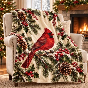 May include: A cream-coloured throw blanket featuring a vibrant red cardinal bird perched on a pine branch. The design includes detailed illustrations of green pine needles, brown pinecones, and clusters of bright red holly berries throughout.