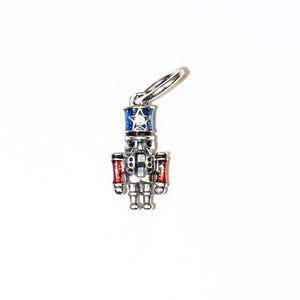 May include: A silver-coloured charm shaped like a nutcracker soldier. The charm features a blue hat with a white star, red arms, and a detailed face. It is attached to a silver ring.