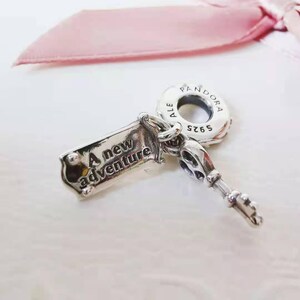 Könnte beinhalten: Ein silberner Charm in Form eines Schlüssels mit der Aufschrift "A new adventure" auf einem rechteckigen Anhänger. Der Schlüssel hat ein detailliertes Design und der Charm ist von der Marke Pandora.