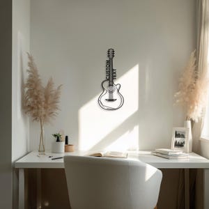 Pode incluir: Uma decoração de parede de guitarra em metal preto com as palavras "CUSTOM TEXT" verticalmente no braço. A guitarra é montada numa parede branca acima de uma secretária. A sala é decorada com capim-dos-pampas e uma foto emoldurada.