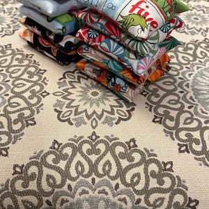 May include: A pile of small, square fabric bean bags with festive patterns. Designs include the Grinch, candy canes, and pumpkins. The bean bags are stacked on a patterned surface.