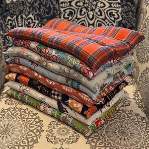 May include: A pile of rectangular pillows with diverse patterns and colours. The top pillow has an orange and blue plaid design. Other pillows display floral, candy cane, and festive prints. The pillows are stacked on a patterned chair.