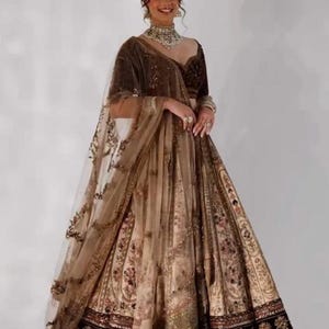 May include: A woman wearing a brown and gold embroidered lehenga. The lehenga features intricate floral patterns and a sheer dupatta. The outfit includes a matching velvet top and jewelry. The text "ZiyaFab" is in the upper right corner.