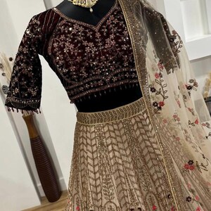 May include: A maroon velvet blouse with intricate floral embroidery and sequin embellishments, paired with a beige skirt featuring a gold geometric pattern. A sheer, floral-printed dupatta completes the ensemble. The text "ZiyaFab" is visible.