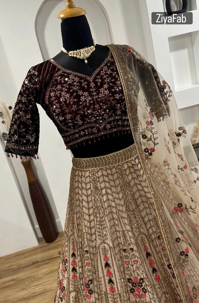 May include: A maroon velvet blouse with intricate floral embroidery and sequin embellishments, paired with a beige skirt featuring a gold geometric pattern. A sheer, floral-printed dupatta completes the ensemble. The text "ZiyaFab" is visible.