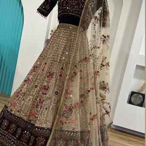 May include: A beige and burgundy Indian lehenga with intricate embroidery and sequin embellishments. The outfit includes a short-sleeved, dark burgundy velvet top and a sheer, floral-patterned dupatta. The text "ZiyaFab" is visible.