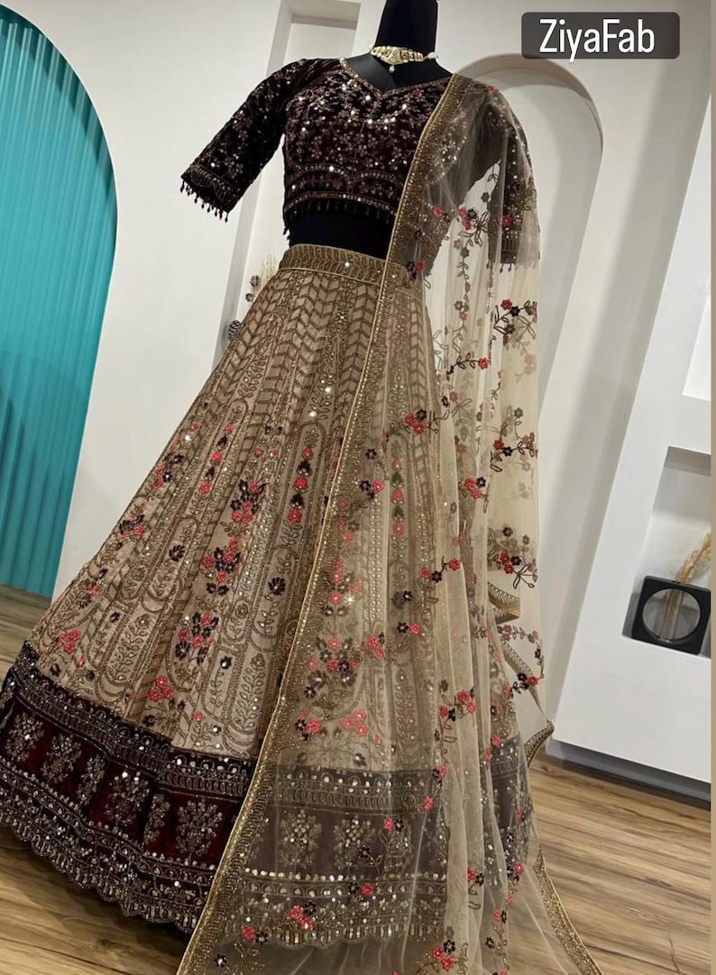 May include: A beige and burgundy Indian lehenga with intricate embroidery and sequin embellishments. The outfit includes a short-sleeved, dark burgundy velvet top and a sheer, floral-patterned dupatta. The text "ZiyaFab" is visible.