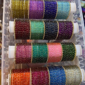 Op de afbeelding: Een collectie veelkleurige glazen armbanden met gouden glitteraccenten, georganiseerd in vier rijen op witte cilindrische houders in een doorzichtige plastic opbergdoos. De armbanden hebben diverse tinten waaronder geel, paars, blauw, rood, groen, roze en zilver, met het label ZiyaFab.