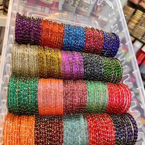 Puede incluir: Un recipiente de almacenamiento de plástico transparente lleno de filas de brazaletes circulares delgados en varios colores, incluyendo morado, naranja, azul, rojo, dorado, verde, rosa y negro. Cada brazalete presenta una superficie texturizada.