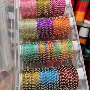 Könnte beinhalten: Ein durchsichtiger Kunststoffbehälter gefüllt mit bunten Glasarmreifen. Die Armreifen sind in Reihen angeordnet und weisen eine Vielzahl von Farben auf, darunter Rosa, Gold, Lila, Orange, Grün, Blau und Rot. Die Armreifen sind mit kleinen, silberfarbenen Verzierungen versehen. Der Text "ZiyaFab" ist sichtbar.