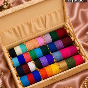 Puede incluir: Una caja de almacenamiento rectangular de color beige que contiene filas de brazaletes finos de colores sólidos en tonos rojo, verde, morado, azul, rosa, negro, blanco, amarillo y dorado. La tapa tiene el texto ZiyaFab en la esquina superior derecha.