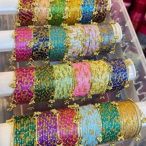 Può includere: Una collezione di braccialetti in metallo multicolore disposti su quattro barre orizzontali bianche all'interno di un contenitore di plastica trasparente. Ogni set presenta una finitura metallica con accenti glitter dorati e piccoli ciondoli pendenti. Il testo ZiyaFab è visibile nell'angolo in alto a destra.