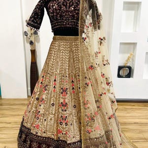 May include: An ornate beige and burgundy lehenga set. The skirt features intricate gold embroidery with floral accents. The dark burgundy velvet blouse has sequin detailing. A sheer dupatta with floral embroidery completes the ensemble. The brand name "ZiyaFab" is visible.