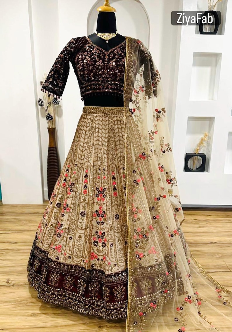 May include: An ornate beige and burgundy lehenga set. The skirt features intricate gold embroidery with floral accents. The dark burgundy velvet blouse has sequin detailing. A sheer dupatta with floral embroidery completes the ensemble. The brand name "ZiyaFab" is visible.